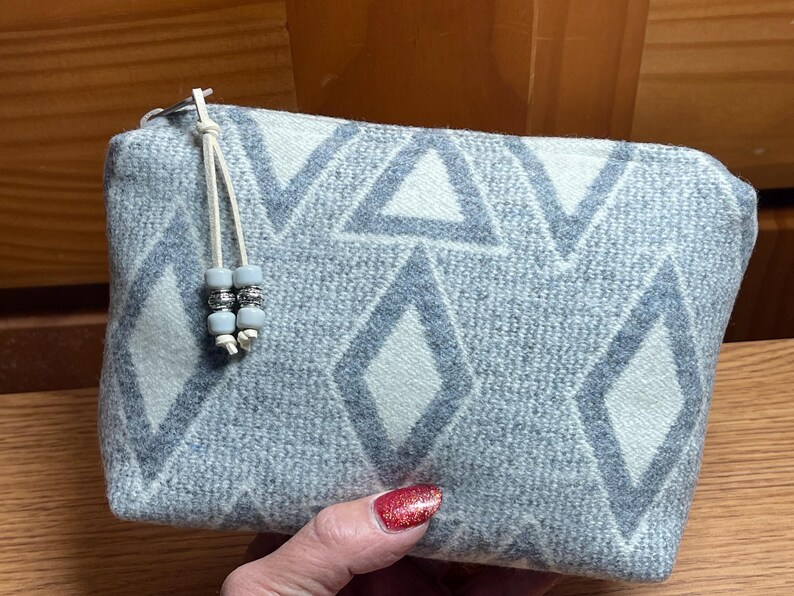 Puede incluir: Un estuche rectangular con un patr&oacute;n geom&eacute;trico azul claro y blanco de diamantes y tri&aacute;ngulos. El estuche tiene un cierre de cremallera y un cord&oacute;n color crema con cuentas decorativas. La tela es suave.