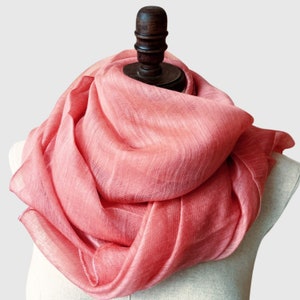 May include: A coral-colored scarf draped over a mannequin. The scarf is made of a lightweight, sheer fabric with a subtle woven texture. The scarf is wrapped around the mannequin's neck and shoulders, creating soft folds and a flowing appearance.