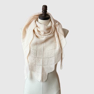 May include: Cream-colored knitted shawl with a geometric pattern and scalloped edges. The shawl is draped over a mannequin, showcasing its design and texture. The soft, neutral color makes it a versatile accessory.