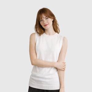 K&ouml;nnte beinhalten: Ein &auml;rmelloser, cremefarbener Pullover mit Rundhalsausschnitt. Das Strickware hat einen gerippten Saum und eine weiche Textur. Das Kleidungsst&uuml;ck wird von einer Person mit rotem Haar und heller Haut getragen. Der Hintergrund ist einfarbig neutral.