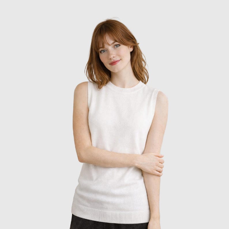 K&ouml;nnte beinhalten: Ein &auml;rmelloser, cremefarbener Pullover mit Rundhalsausschnitt. Das Strickware hat einen gerippten Saum und eine weiche Textur. Das Kleidungsst&uuml;ck wird von einer Person mit rotem Haar und heller Haut getragen. Der Hintergrund ist einfarbig neutral.