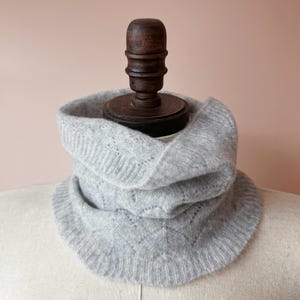 May include: A gray knitted cowl neck scarf with a delicate openwork pattern. The scarf is displayed on a mannequin.