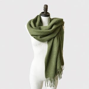 May include: An olive green scarf with a soft, textured appearance draped around a white mannequin. The scarf features a long, rectangular shape with fringe detailing along the bottom edges, providing a simple and functional design for neck coverage.