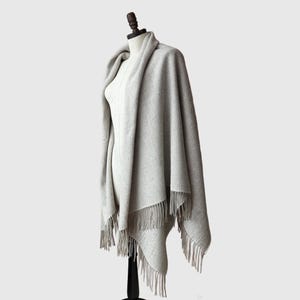 Puede incluir: Un chal gris claro con estampado de espiga y flecos. El chal está drapeado sobre un maniquí, mostrando su diseño y textura. Los flecos cuelgan a lo largo del borde inferior del chal.
