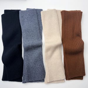 May include: Four pairs of ribbed knit leg warmers in a variety of colors: navy blue, gray-blue, cream, and brown. The leg warmers are folded and displayed on a white background. The knitwear has a vertical rib pattern.