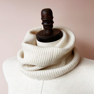 Puede incluir: Una bufanda de cuello de tortuga de punto de color crema. La bufanda es de punto de canalé y está doblada sobre sí misma. La bufanda se muestra en un maniquí blanco.