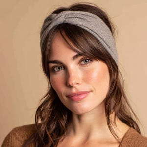 May include: A gray headband with a twisted knot design. The headband is made of a soft, knitted material. The model has brown hair and is wearing a brown sweater.