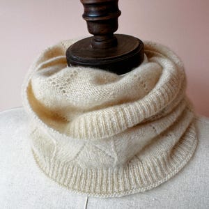 May include: A cream-colored knitted infinity scarf with a delicate openwork pattern. The scarf is folded and displayed on a mannequin.