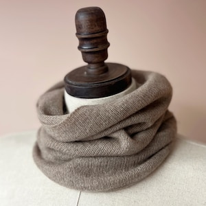 May include: A brown knitted neck warmer or cowl on a mannequin. The neck warmer is made of a soft, textured fabric.