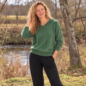 May include: A sage green long-sleeve hooded jumper with a V-neckline and matching drawstring cords, paired with solid black trousers. The jumper features ribbed cuffs and a ribbed hem, providing a clean, structured look for the garment.