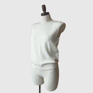 K&ouml;nnte beinhalten: Cremefarbener &auml;rmelloser Pullover auf einer Schaufensterpuppe. Der Pullover hat einen gerippten Saum und einen Rundhalsausschnitt. Die Schaufensterpuppe ist hellbeige und hat einen Holzhals. Der Hintergrund ist einfarbig hellgrau.