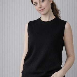 May include: A black sleeveless sweater with a crew neck. The sweater is made of a soft material and is worn with black pants. The background is a light gray wall with vertical lines.