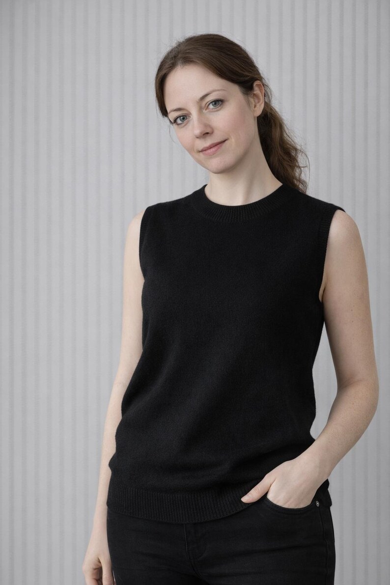 May include: A black sleeveless sweater with a crew neck. The sweater is made of a soft material and is worn with black pants. The background is a light gray wall with vertical lines.