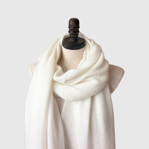 May include: A cream-colored scarf draped over a mannequin. The scarf is made of a soft, woven material and is wrapped around the mannequin's neck and shoulders. The scarf is a solid color and appears to be lightweight.