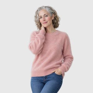 May include: A soft, fuzzy, long-sleeved jumper in a muted dusty pink colour with a classic crew neckline and ribbed cuffs, paired with blue denim jeans. The jumper features a textured, brushed knit finish and a relaxed, comfortable fit.