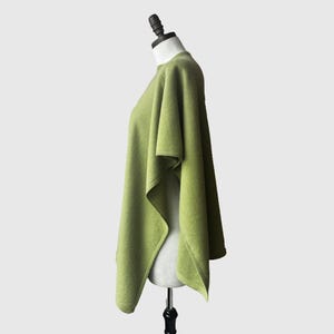 May include: A green poncho made of soft fabric. The poncho is draped over a mannequin and has a simple, elegant design.