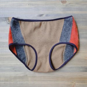 May include: High-waisted underwear featuring a color-blocked design with a central tan panel, dark gray side panels, and vibrant orange outer edges. The garment is finished with dark navy blue trim along the waistband and leg openings.