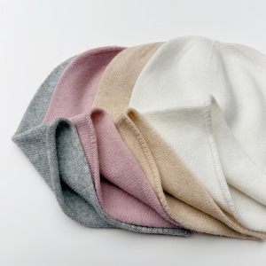 May include: A collection of five knitted beanies in various colors: gray, pink, beige, and white. The beanies are neatly stacked, showcasing their soft texture and simple design. The hats are made of a fine knit material.