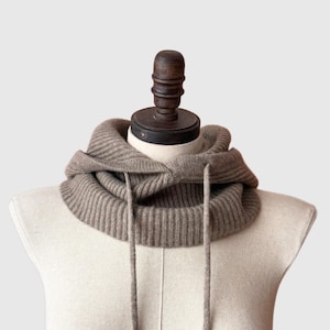 May include: A taupe-colored hooded cowl neck warmer made of ribbed knit fabric. The cowl features a drawstring and is displayed on a mannequin. The neck warmer is designed to be worn around the neck and over the head.