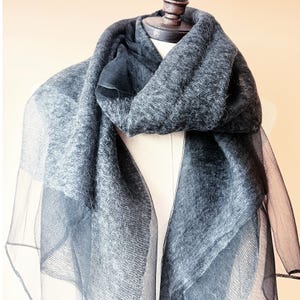 May include: A dark gray scarf draped over a mannequin. The scarf has a textured, woven appearance with a sheer, darker gray border. The scarf is made of a soft material and is designed to be worn around the neck.