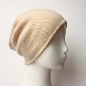 May include: A cream-colored beanie hat made of a soft, knitted material. The hat is displayed on a mannequin head, showcasing its shape and texture. The hat has a ribbed brim and a slightly slouchy fit.