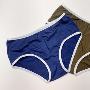 May include: A pair of high-waisted briefs in solid blue and olive green, both featuring a contrasting white trim along the waistband and leg openings. The fabric appears soft and lightweight, with a simple design focused on comfort and coverage.