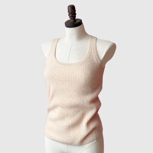 May include: A light peach-colored ribbed tank top displayed on a mannequin. The tank top has a scoop neck and a fitted silhouette. The fabric appears to be soft and has vertical ribbing throughout the garment. The mannequin has a wooden neck and shoulder piece.