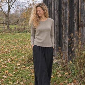 May include: A long-sleeved, crew-neck sweater in a heathered beige tone paired with loose-fitting, ankle-length black trousers featuring side pockets. The sweater has a ribbed hem and cuffs, while the trousers have a simple, straight-leg silhouette.