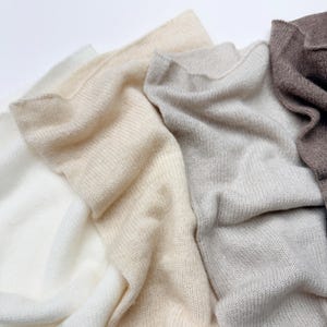 May include: Close-up of folded knitwear in shades of white, beige, light gray, and brown. The soft, textured fabric is arranged in overlapping layers, showcasing the different colors and textures of the material. The knitwear appears to be made of a soft, possibly natural fiber.
