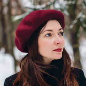 May include: A woman wearing a burgundy beret and a black turtleneck sweater. She is standing in a snowy forest.