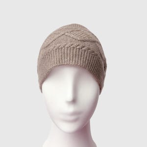 May include: A taupe-colored knit beanie hat with a cable knit design on the crown. The hat is displayed on a white mannequin head. The beanie has a ribbed brim and a textured pattern on the top.