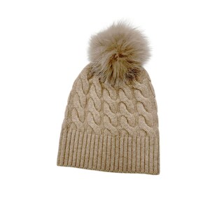 May include: A beige knitted beanie with a fluffy brown pom pom on top. The beanie has a cable knit pattern.