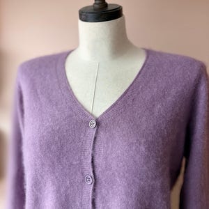 May include: A lavender-colored cardigan sweater with a V-neck and three matching buttons. The sweater is displayed on a mannequin. The soft, fuzzy texture of the knitwear is visible.