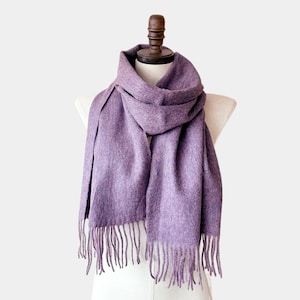 May include: A solid lavender scarf with a soft, fuzzy texture draped around a mannequin neck. The scarf features a rectangular shape with long, straight fringe tassels along the bottom edges, creating a simple and uniform appearance in light purple.