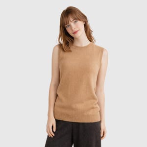 May include: A sleeveless, tan-colored sweater. The sweater has a crew neck and a ribbed hem. The model is wearing dark gray pants. The sweater is made of a soft, knit material. The overall style is casual and comfortable.