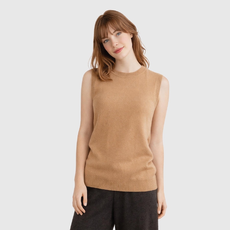 May include: A sleeveless, tan-colored sweater. The sweater has a crew neck and a ribbed hem. The model is wearing dark gray pants. The sweater is made of a soft, knit material. The overall style is casual and comfortable.