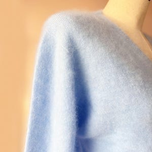 May include: A light blue, fluffy sweater with a V-neck design. The soft knitwear is displayed on a mannequin, showcasing the texture and color of the garment. The sweater's delicate fabric suggests a cozy and comfortable feel.