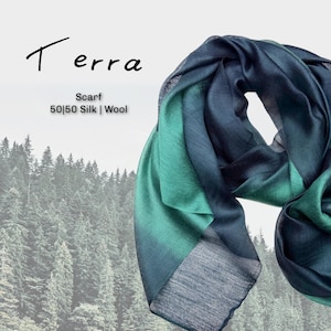 May include: A draped scarf in shades of teal and navy blue, with the word "Terra" in cursive script. The scarf is made of a blend of 50% silk and 50% wool. The background features a blurred image of a forest.