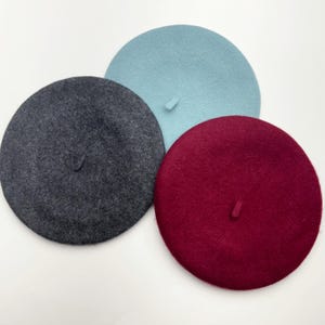 May include: Three wool berets in different colors: dark gray, light blue, and burgundy. Each beret has a small, rounded stem on top. The berets are arranged on a white surface.