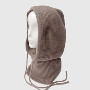 May include: A taupe-colored knitted balaclava with a ribbed texture. The hood features a drawstring closure and is displayed on a mannequin head. This winter accessory provides warmth and style.