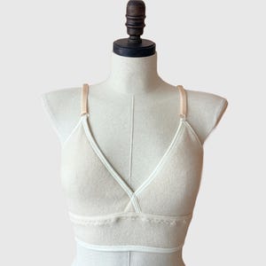 May include: A cream-colored bralette with a V-neck design. The bra features thin, light-colored straps and a decorative trim along the bottom edge. The bralette is displayed on a mannequin.