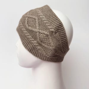 May include: A taupe-colored knitted headband with a cable knit design. The headband is displayed on a white mannequin head. The cable knit pattern is centered on the front of the headband, with a textured diamond shape.