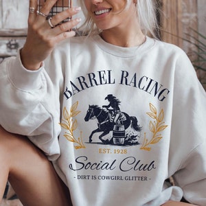 Pu&ograve; includere: Felpa bianca con una grafica di una cowgirl che cavalca un cavallo intorno a una botte. La grafica dice "Barrel Racing Social Club - Dirt is Cowgirl Glitter - Est. 1928".