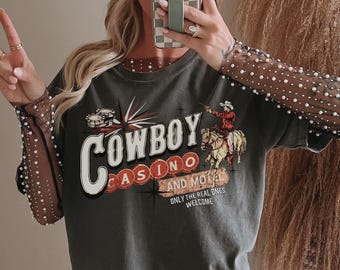 Peut inclure: Un t-shirt gris foncé avec un motif vintage d'un cow-boy à cheval devant un panneau qui indique "Cowboy Casino and Motel". Le panneau a une étoile rouge et les mots "Only the real ones welcome". Le t-shirt a un col rond et des manches courtes.