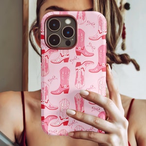May include: Pink phone case with a repeating pattern of pink cowboy boots and bows. The case has a red border around the camera and edges. The boots have intricate details and are set against a light pink background.