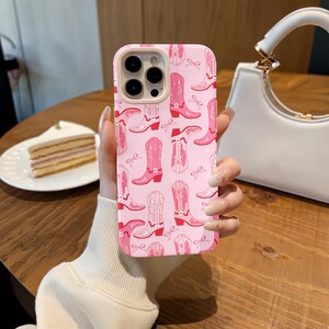May include: Pink phone case with a repeating pattern of pink and red cowboy boots and bows. The case has a light pink border around the camera lenses. The phone case is held in a person's hand.