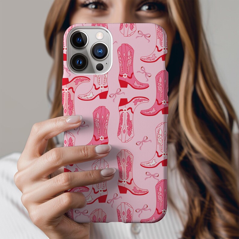 May include: A pink phone case featuring a repeating pattern of pink cowboy boots and red bows. The boots have decorative stitching and small floral accents. The case is held in front of a person.