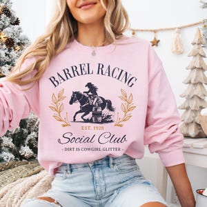 Pu&ograve; includere: Felpa rosa con un design grafico di una cowgirl a cavallo e un barile. Il testo recita "Barrel Racing Social Club - Dirt is Cowgirl Glitter - Est. 1928".