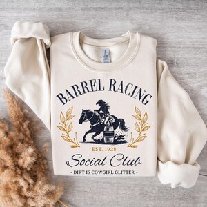 Pu&ograve; includere: Una felpa color crema con un design grafico blu navy. La grafica presenta una cowgirl che cavalca un cavallo intorno a una botte. Il testo recita "Barrel Racing Social Club Est. 1928 - Dirt is Cowgirl Glitter - "