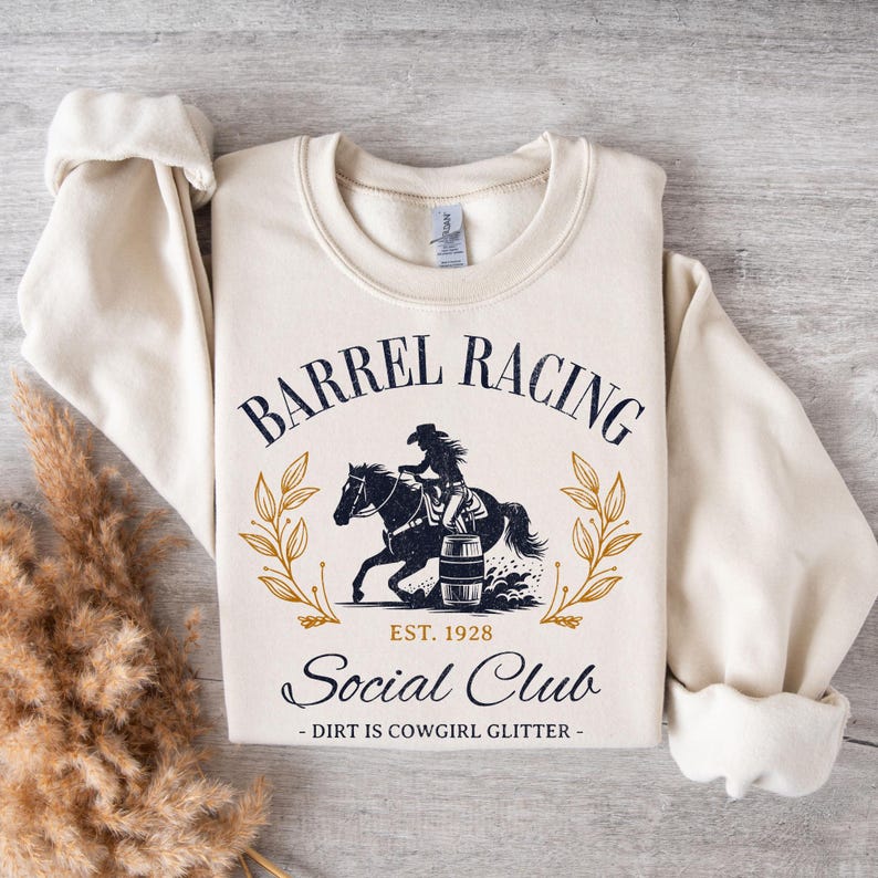 Pu&ograve; includere: Una felpa color crema con un design grafico blu navy. La grafica presenta una cowgirl che cavalca un cavallo intorno a una botte. Il testo recita "Barrel Racing Social Club Est. 1928 - Dirt is Cowgirl Glitter - "